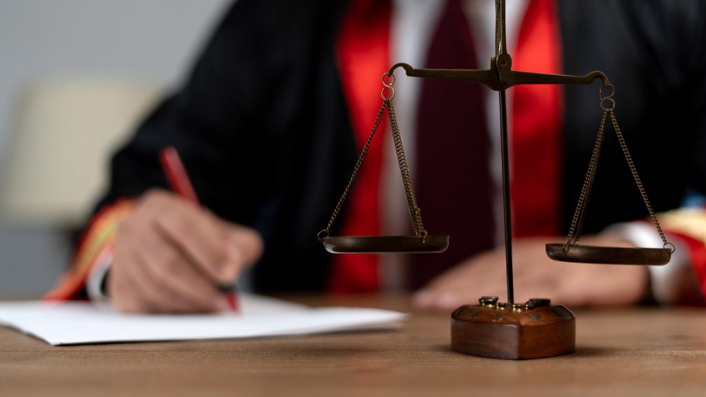 A judge, dressed in a black and red robe, writes on a piece of paper at a wooden desk. In the foreground, a brass balance scale is in focus, symbolizing justice in contesting wills.
