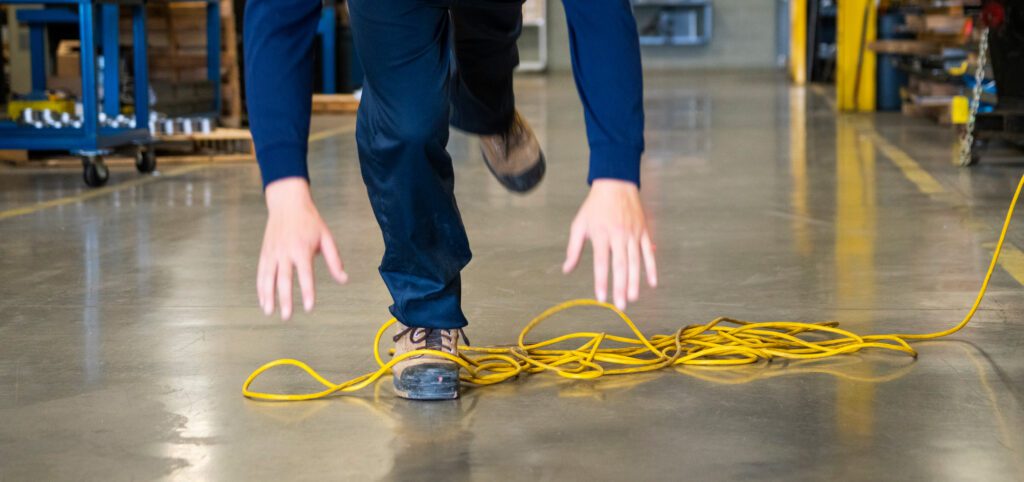 A person in a warehouse is experiencing a trip and fall accident, stumbling over a yellow extension cord on the floor with arms extended for balance.