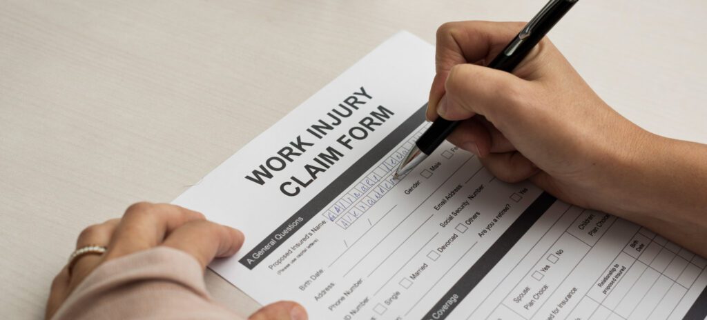 A person fills out a "Work Injury Claim Form" with a pen on a table, preparing to provide information about an injury for workplace compensation or a potential Workplace Injury Lawsuit.