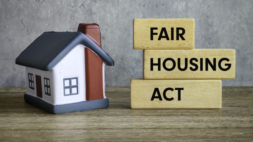 A small model house sits on a wooden surface next to three stacked wooden blocks with "Fair Housing Act" written in black letters, highlighting protections against housing discrimination and disability. The background is a plain gray wall.