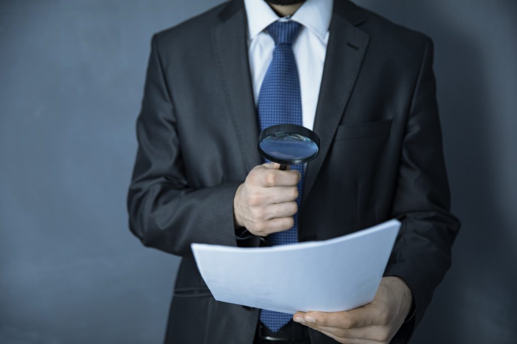 A hand holding a magnifying glass over a document, representing the review of cremation logs and chain-of-custody records in a mortuary negligence case.