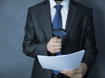 A hand holding a magnifying glass over a document, representing the review of cremation logs and chain-of-custody records in a mortuary negligence case.
