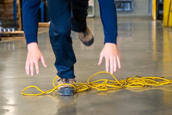 A person in a warehouse is experiencing a trip and fall accident, stumbling over a yellow extension cord on the floor with arms extended for balance.