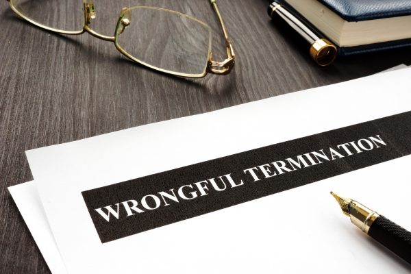 A document titled "Wrongful Termination" lies on a desk next to a pair of eyeglasses, a blue notebook, and a fountain pen, suggesting what to do if fired or wrongfully terminated is under careful consideration.
