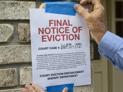 A person tapes a "Final Notice of Eviction" with a court case number to a brick wall near a door, highlighting concerns about baseless evictions, harassment, and retaliation from the court eviction enforcement sheriff department.
