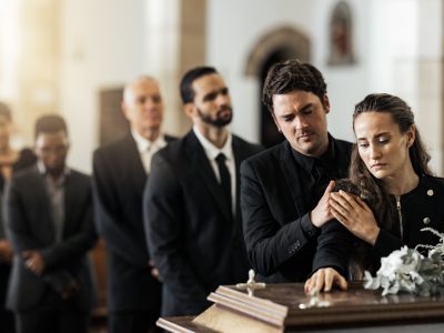 Mourners in black clothing stand by a casket adorned with white flowers. As a man comforts a grieving woman, it's important to know your rights in cases of negligent mishandling of human remains. Others stand solemnly in the background.
