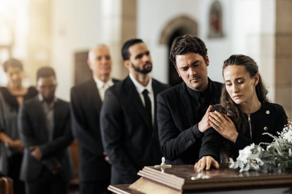 Mourners in black clothing stand by a casket adorned with white flowers. As a man comforts a grieving woman, it's important to know your rights in cases of negligent mishandling of human remains. Others stand solemnly in the background.