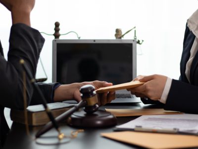 Two people prepare for a deposition in suits sit at a desk with a gavel, scales of justice, and a laptop. One hands an envelope to the other, suggesting deposition preparation or guidance on how to prepare for a deposition during a legal meeting.