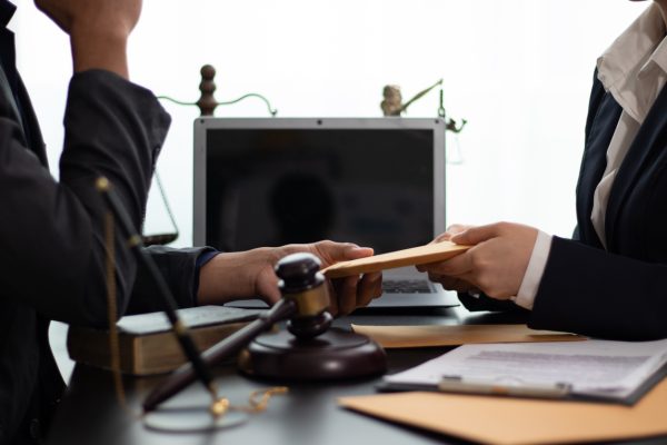 Two people prepare for a deposition in suits sit at a desk with a gavel, scales of justice, and a laptop. One hands an envelope to the other, suggesting deposition preparation or guidance on how to prepare for a deposition during a legal meeting.