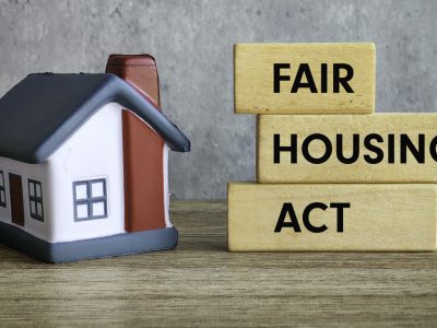 A small model house sits on a wooden surface next to three stacked wooden blocks with "Fair Housing Act" written in black letters, highlighting protections against housing discrimination and disability. The background is a plain gray wall.