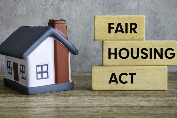 A small model house sits on a wooden surface next to three stacked wooden blocks with "Fair Housing Act" written in black letters, highlighting protections against housing discrimination and disability. The background is a plain gray wall.