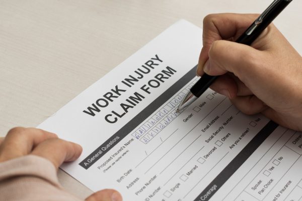 A person fills out a "Work Injury Claim Form" with a pen on a table, preparing to provide information about an injury for workplace compensation or a potential Workplace Injury Lawsuit.