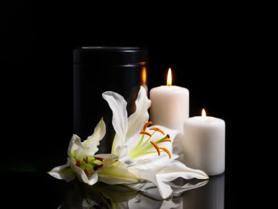 A mortuary urn beside burning candles and white lily flowers, representing families harmed by the mishandling of human remains in California.