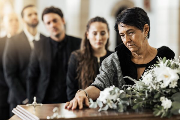 A woman stands by a coffin, resting her hand on it, engulfed in the somber atmosphere of mourning. As she reflects on the possibility of the Mishandling of Human Remains, white flowers adorn the coffin, reminiscent of past developments. In the background, black-clad mourners share her sorrowful vigil.