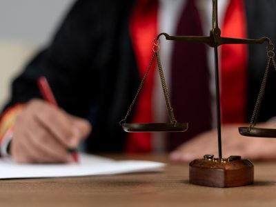 A judge, dressed in a black and red robe, writes on a piece of paper at a wooden desk. In the foreground, a brass balance scale is in focus, symbolizing justice in contesting wills.
