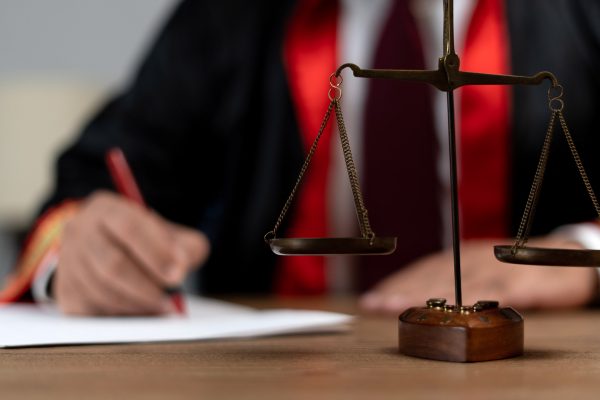 A judge, dressed in a black and red robe, writes on a piece of paper at a wooden desk. In the foreground, a brass balance scale is in focus, symbolizing justice in contesting wills.
